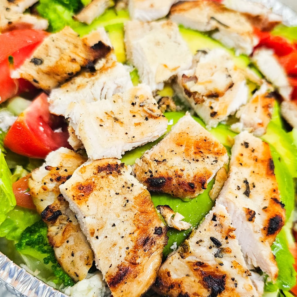 Grilled chicken pieces served over rice with lettuce, tomatoes, broccoli, and green peppers in a foil tray.