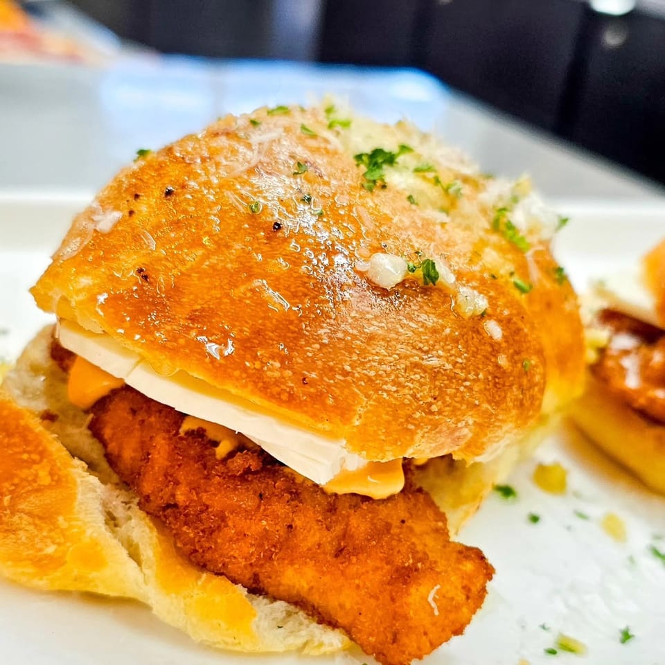 Close-up of a crispy fried chicken sandwich on a toasted herb-topped bun with cheese, served on a white plate.