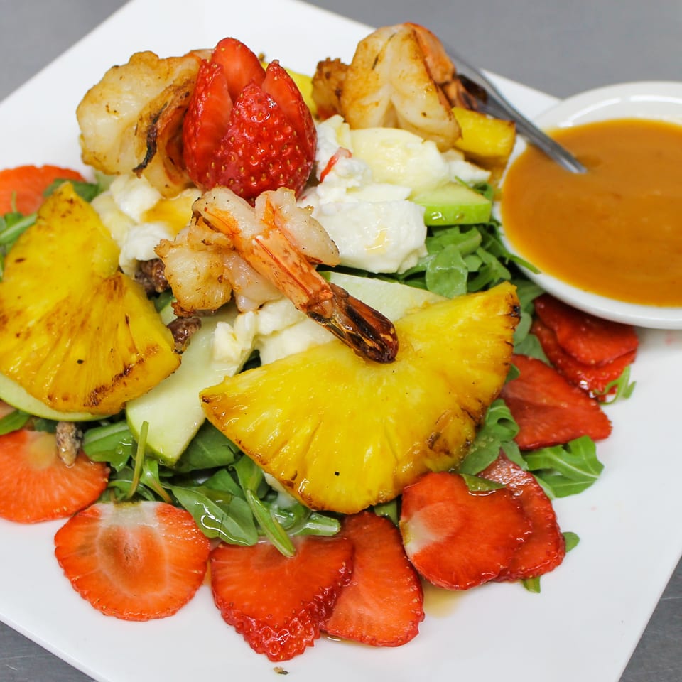 Salad topped with grilled shrimp, pineapple, strawberries, cheese, greens, and roasted vegetables, served with orange dressing.